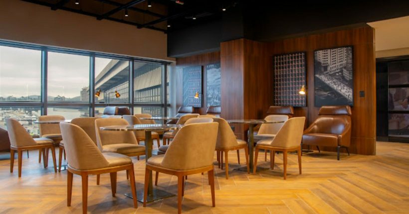 Interior da segunda sala VIP do W Premium no Aeroporto de Guarulhos. Este espaço oferece um buffet self-service, bar com opções alcoólicas e não alcoólicas, ducha, serviço de barista e uma vista panorâmica para o pátio de aeronaves.